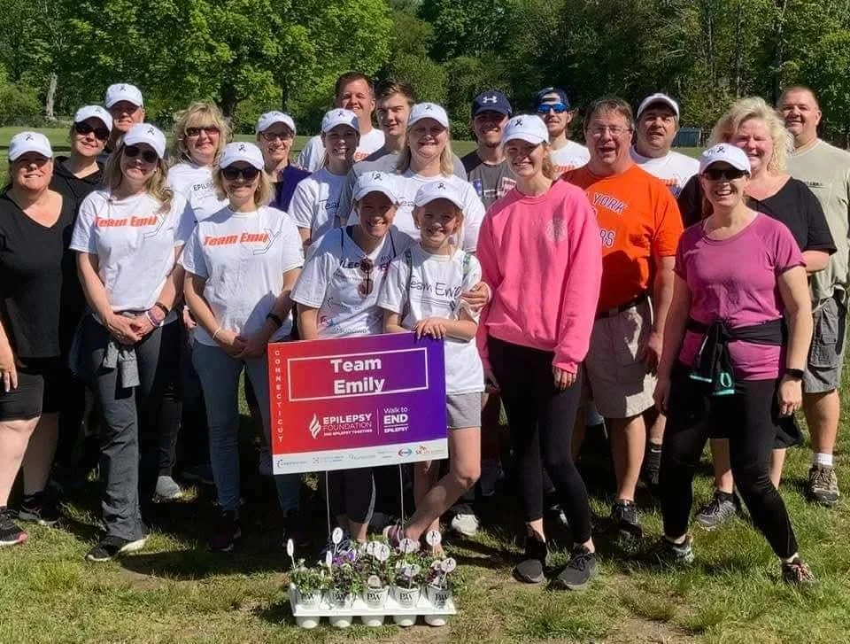Group of smiling people, including children and adults, posing outdoors on a sunny day at a walk event supporting epilepsy awareness. They are holding a sign that says 'Team Emily' and standing behind potted flowers.