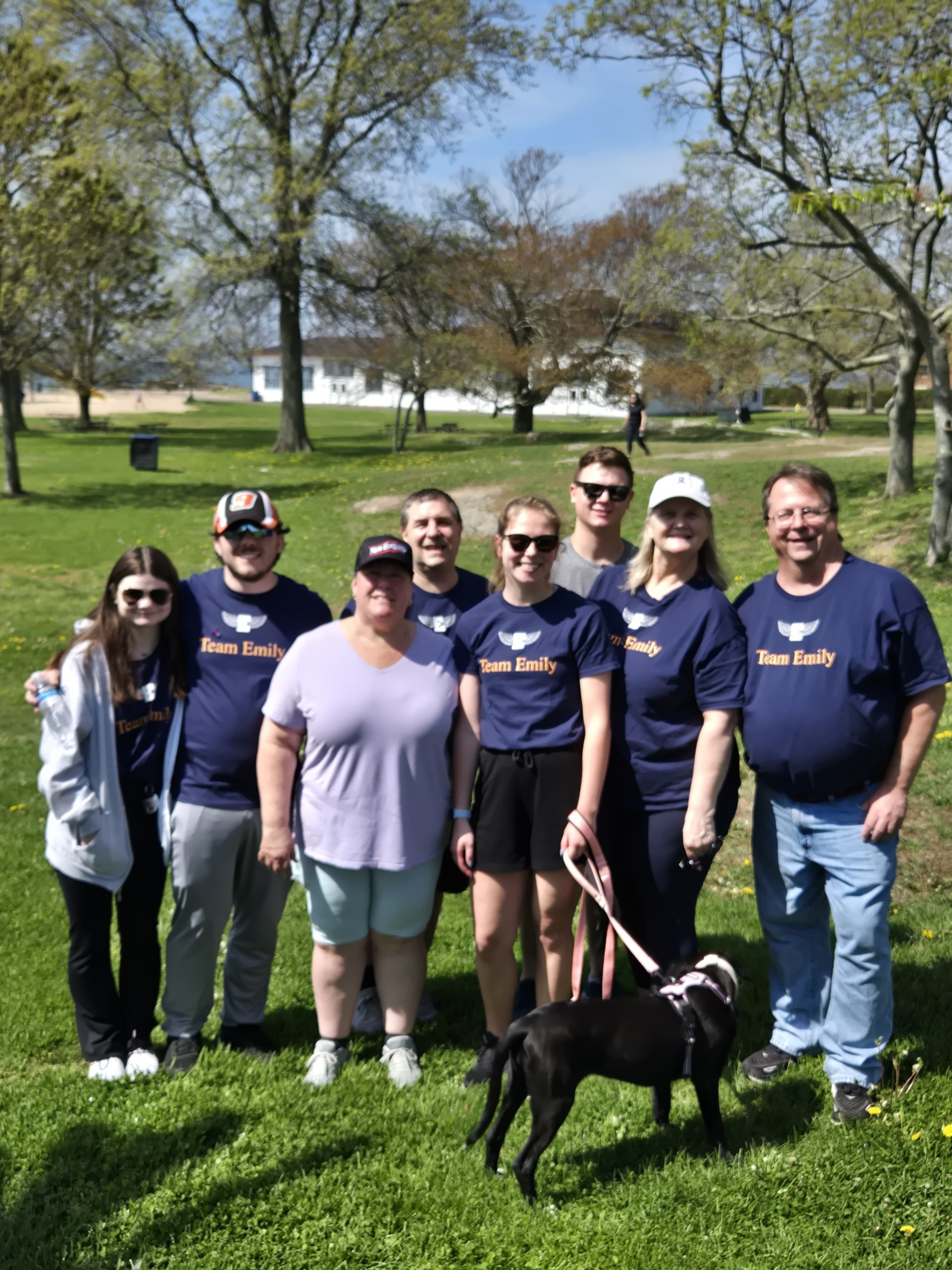 Group of eight people standing outdoors on grass with trees and blue sky in the background, one person holding a small dog.