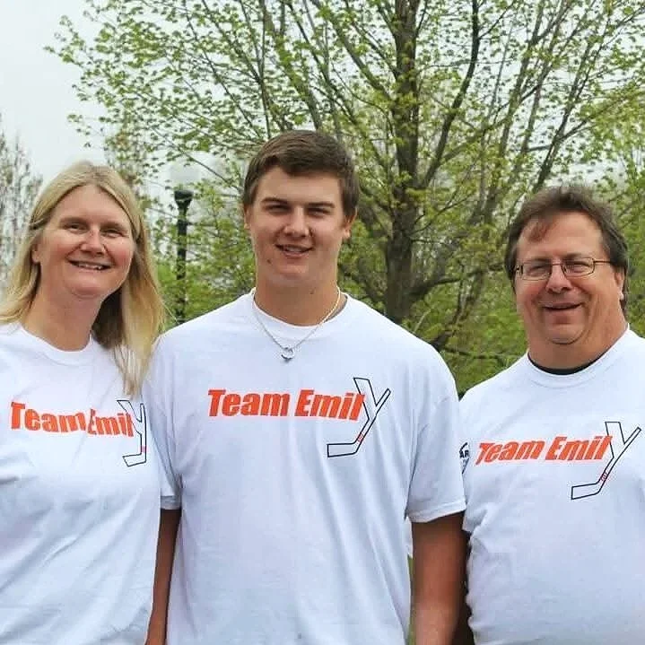 Three people standing outdoors wearing white t-shirts with Team Emily written on it, and a hockey stick for the Emily Yish Remembrance Fund.