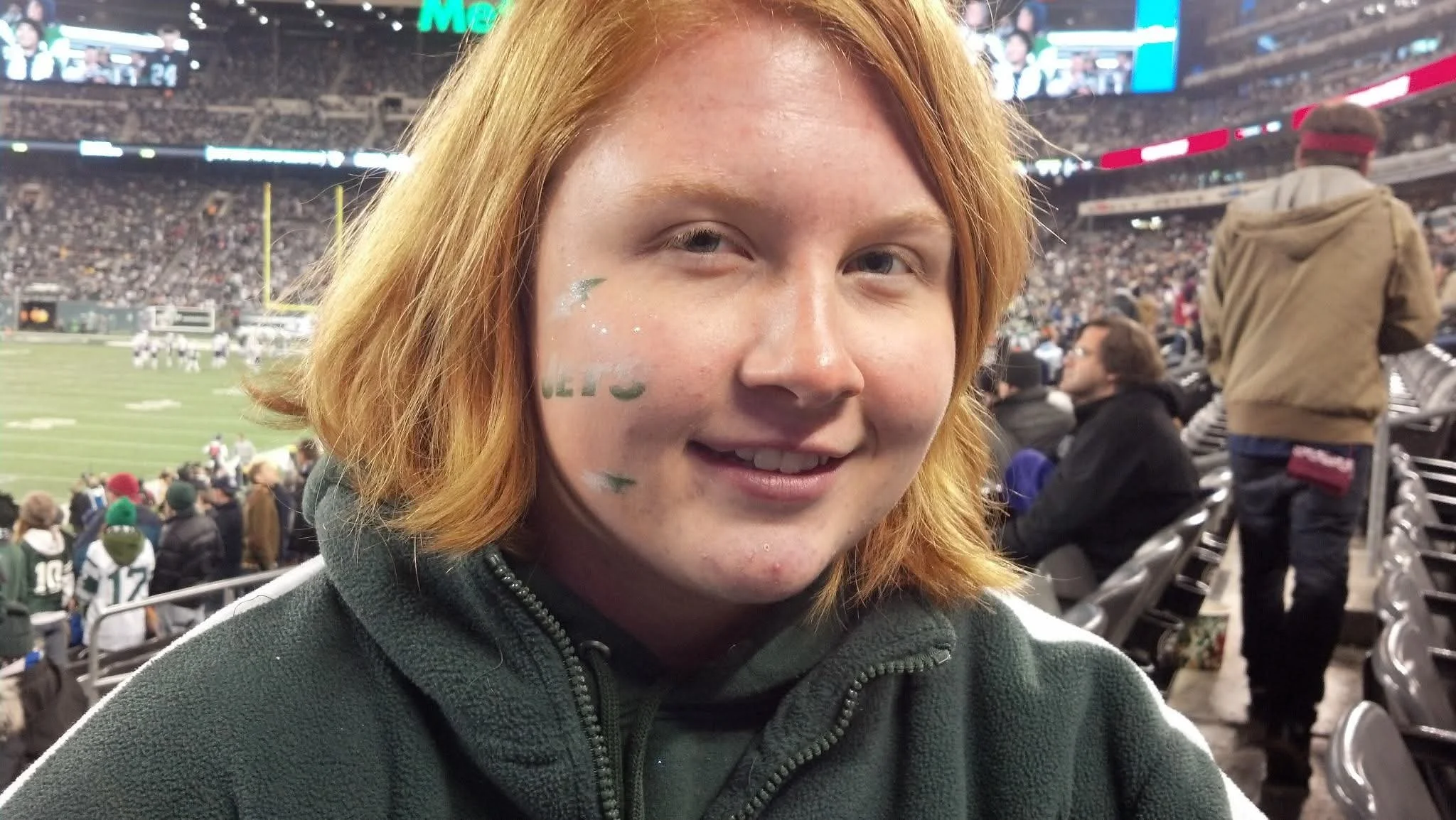 A young woman with red hair smiling at a football game in a stadium. She has green face paint and glitter on her cheek, and is wearing a dark gray zip-up jacket. The background shows a crowded stadium with people seated and standing, and a football field with players.