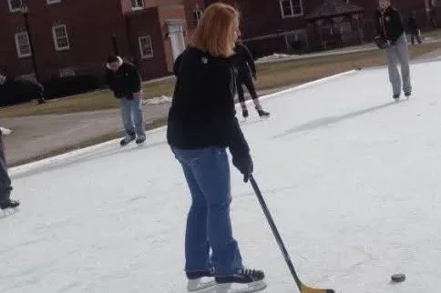 Person in black coat and blue jeans playing ice hockey on an outdoor ice rink, using a hockey stick and puck, with several other people on the ice and brick buildings in the background.