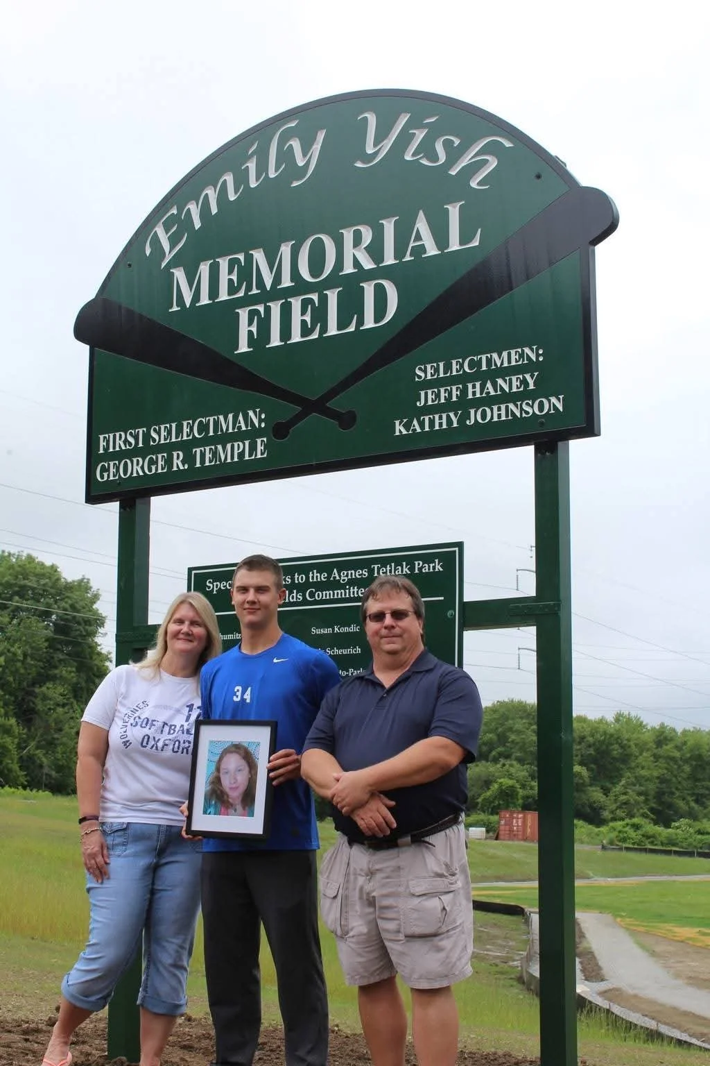 Three people standing together at a memorial field sign, holding a framed photograph of a young woman with long red hair. The sign reads 'Family Wish Memorial Field' and mentions first and selectmen names, with a green background and white text.