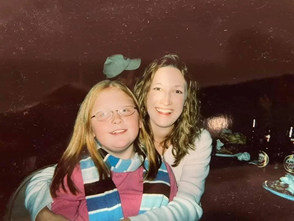 A woman and a young girl sitting together, smiling and hugging indoors with a dark wall behind them.