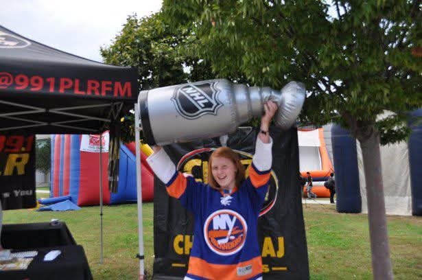 A woman wearing a New York Islanders hockey jersey holding a large silver NHL trophy replica above her head outdoors.