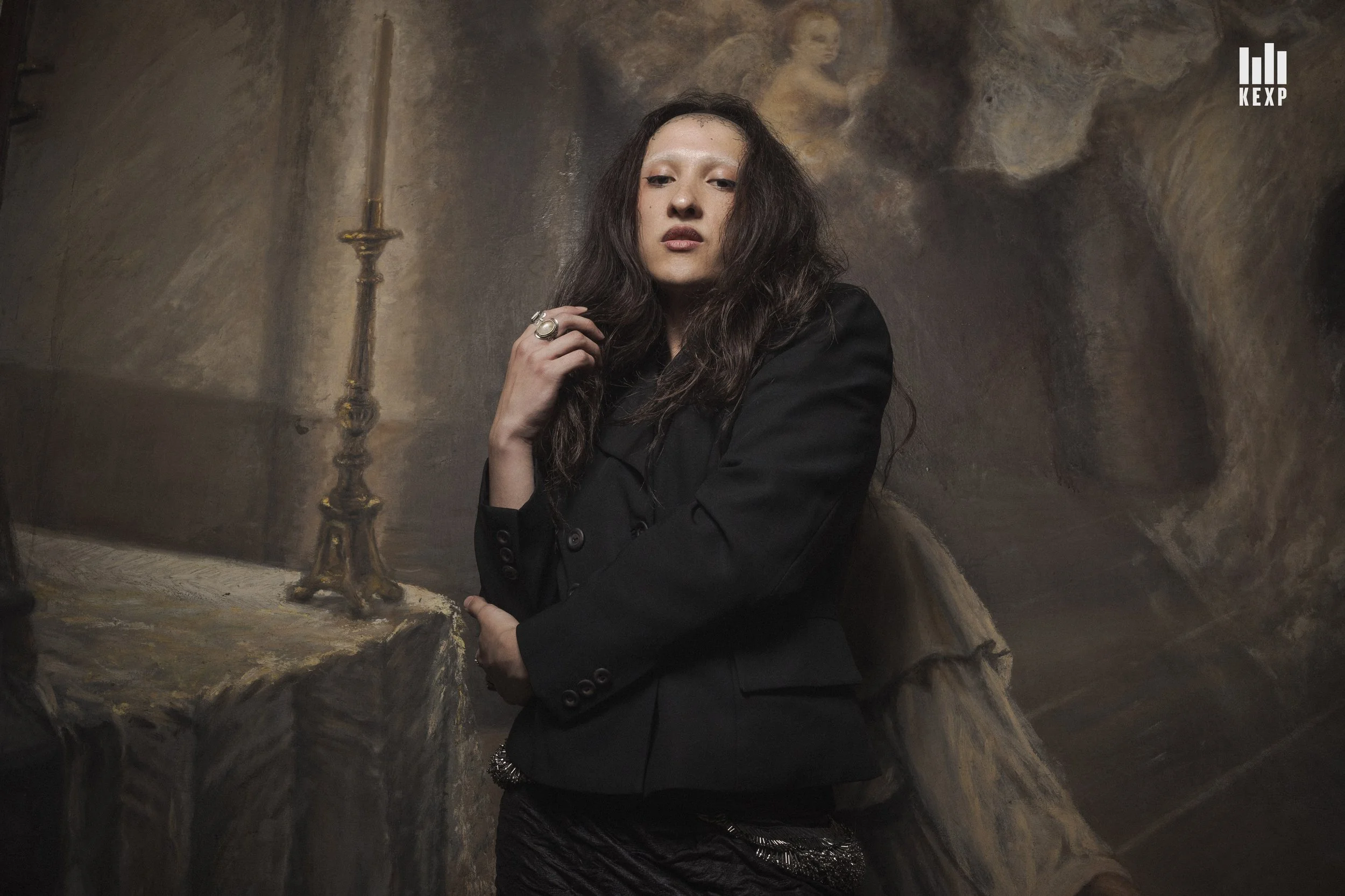 A woman with dark, wavy hair and light makeup stands in front of a mural, wearing a black blazer and rings on her fingers.