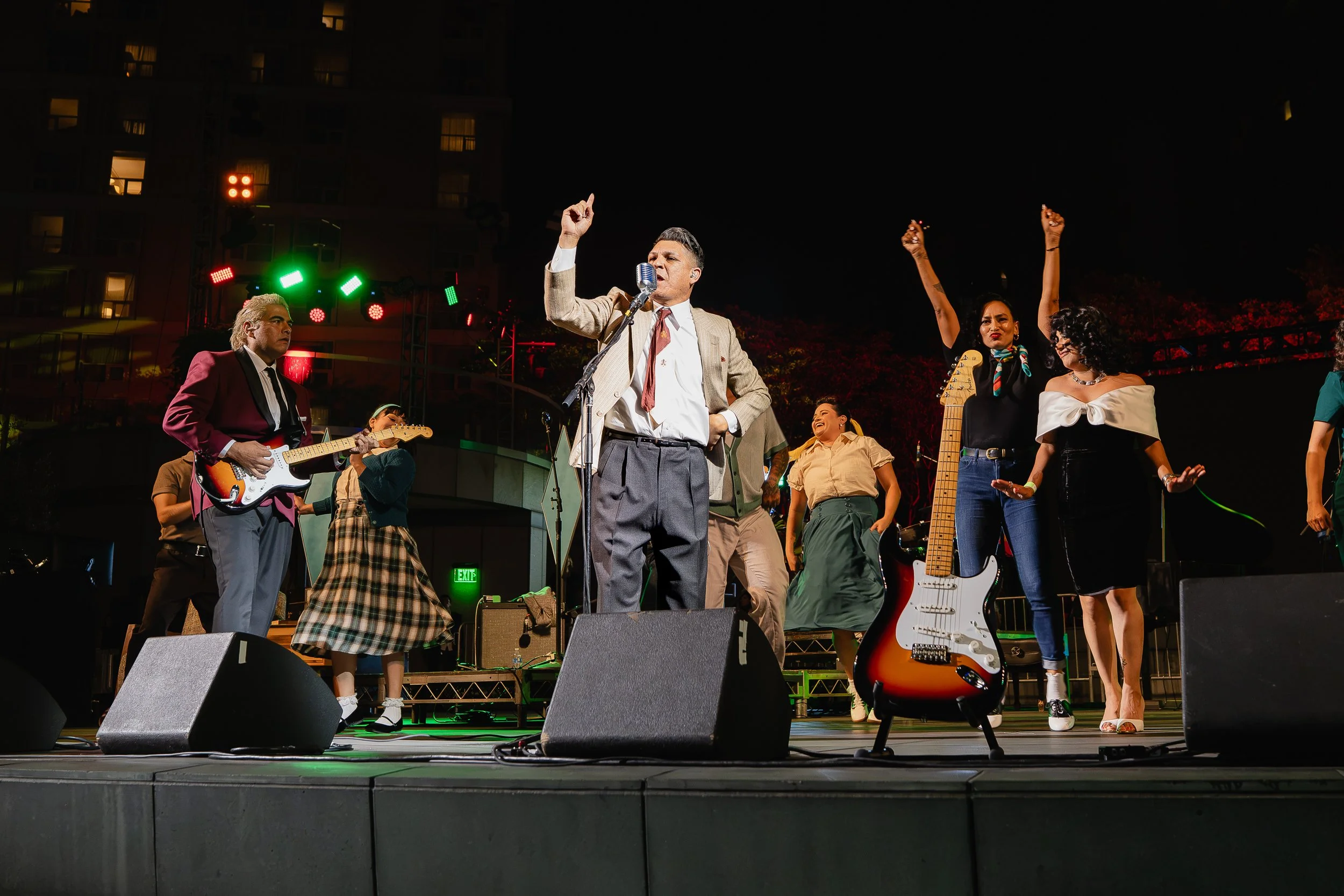 People performing on an outdoor stage at night. The lead singer, dressed in a beige suit, is singing into a vintage microphone, with several other musicians playing guitars and dancing around him. The stage is lit with colorful lights.