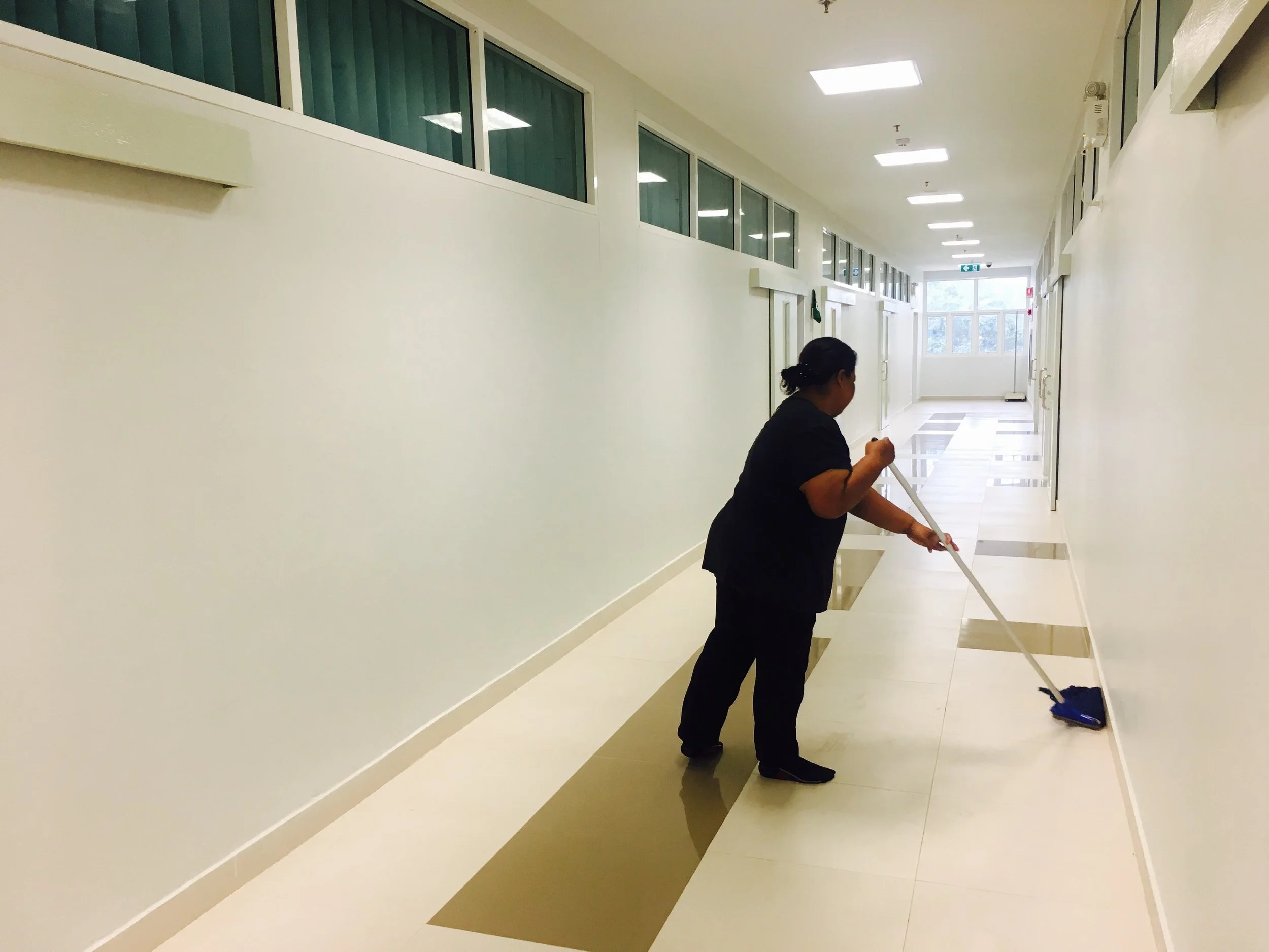 A woman in black uniform cleaning the floor of a corridor with a mop in a modern building with white walls and large windows