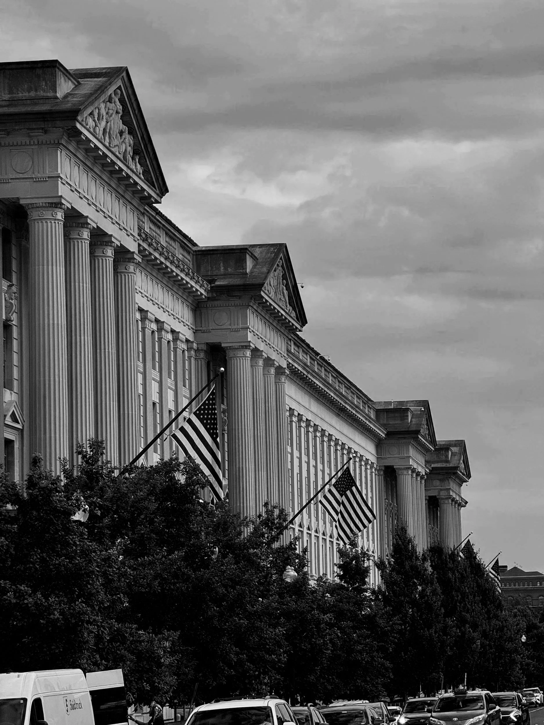 Black and white photograph of the White House with American flags hanging outside, trees, parked cars in front, and a cloudy sky.