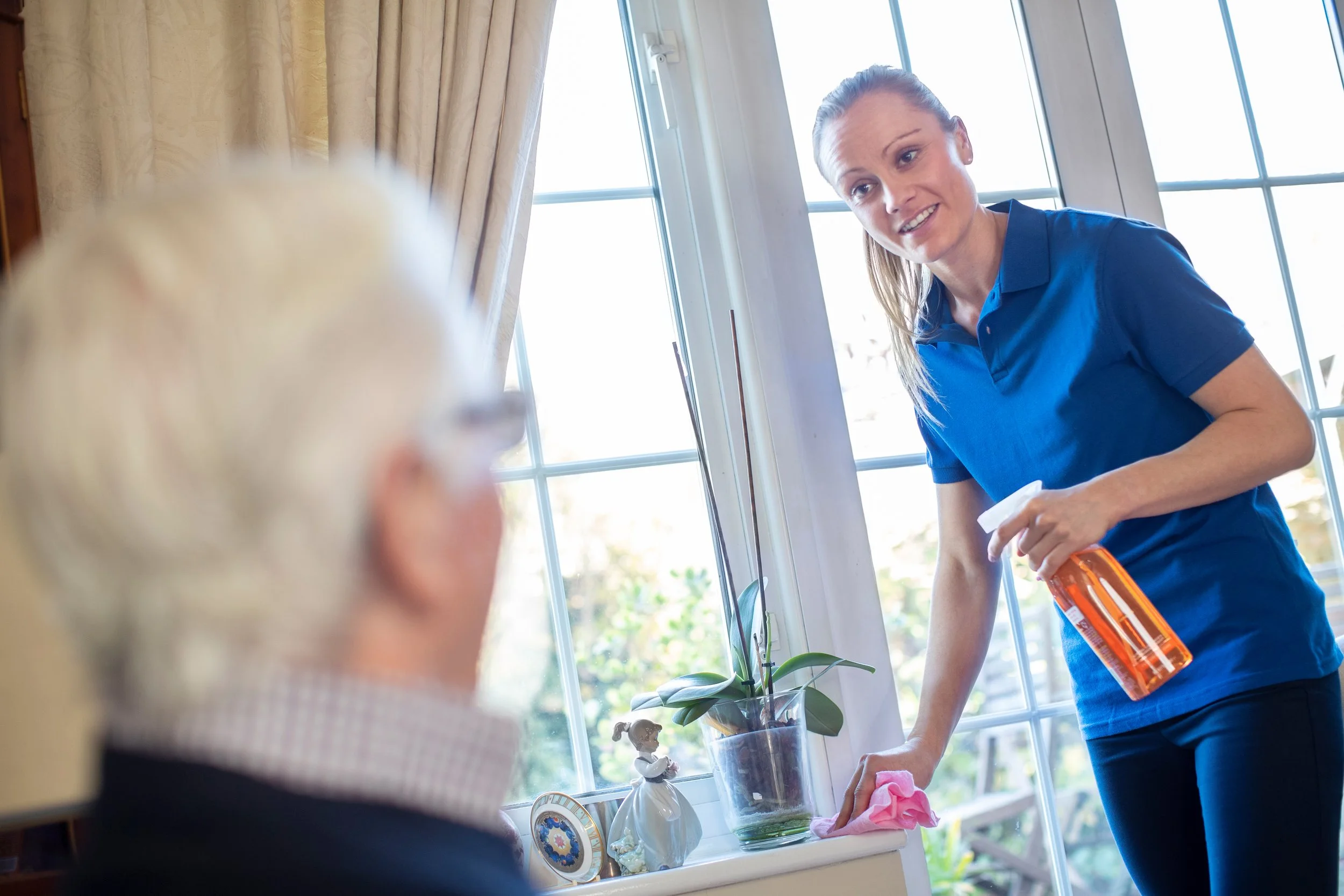 A young woman in a blue polo shirt mops or cleans a windowsill with a pink cloth, holding a spray bottle, while talking to an older woman with white hair in glasses, sitting nearby. Sunlight streams through large windows, and a potted plant and decorative items are on the windowsill.