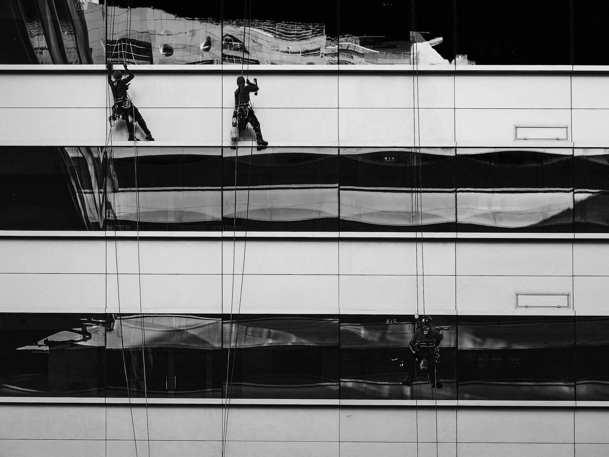 Three window washers cleaning the exterior of a multi-story glass building, reflected in the glass windows.