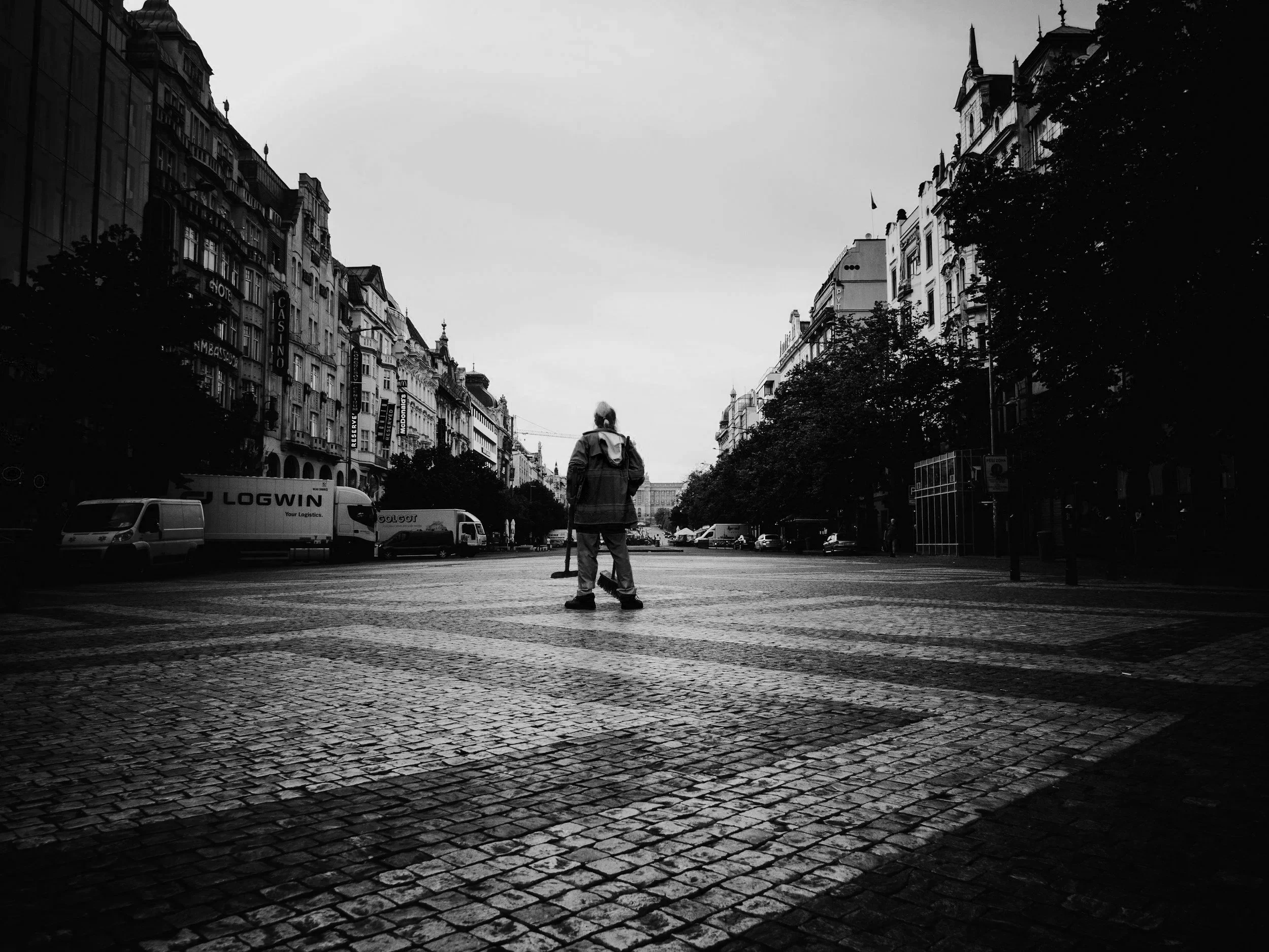 A person standing in the middle of a cobblestone street in an urban area, seen from behind, with tall historic buildings and trees lining the street, and vehicles parked along the sides.