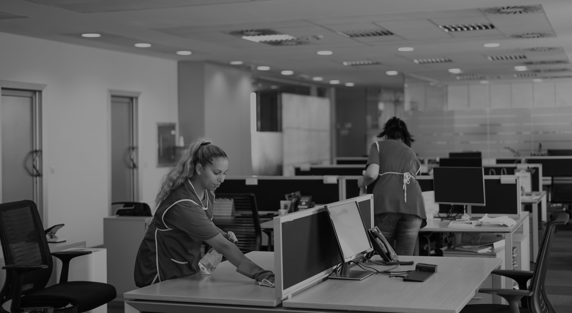 Two women working in an office, one with long hair and another with short hair, organizing papers and cleaning around computer desks.