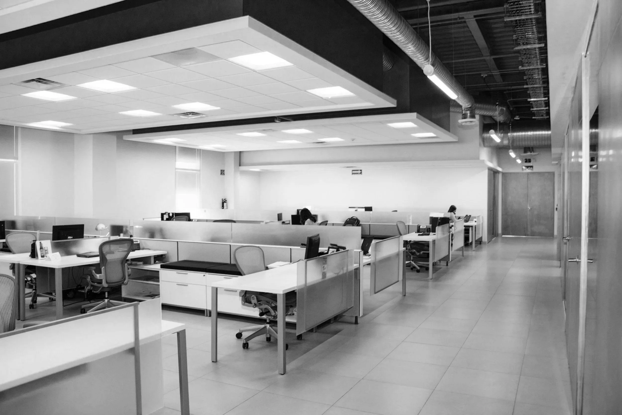 Empty office with desks, chairs, and workstations, with a woman working at a desk in the background.