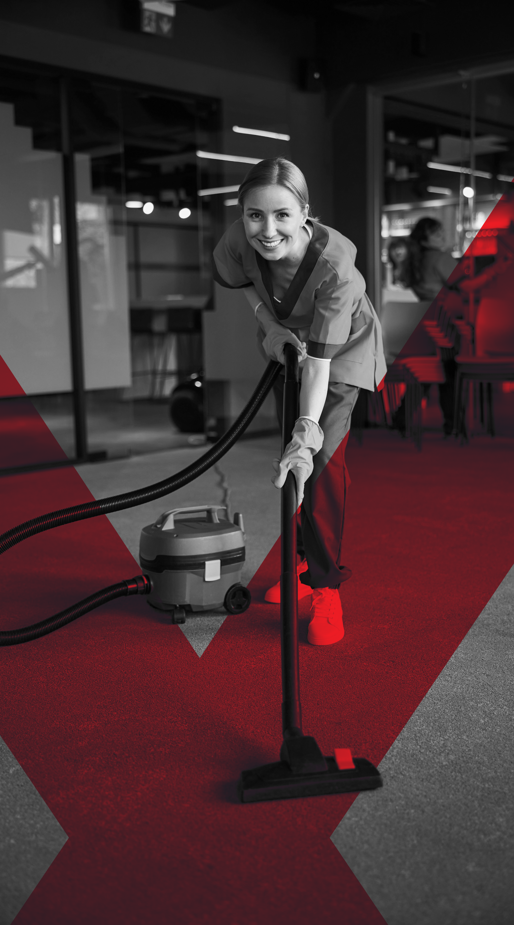 A woman in medical scrubs vacuuming a carpeted floor in an office or hospital setting, smiling at the camera.