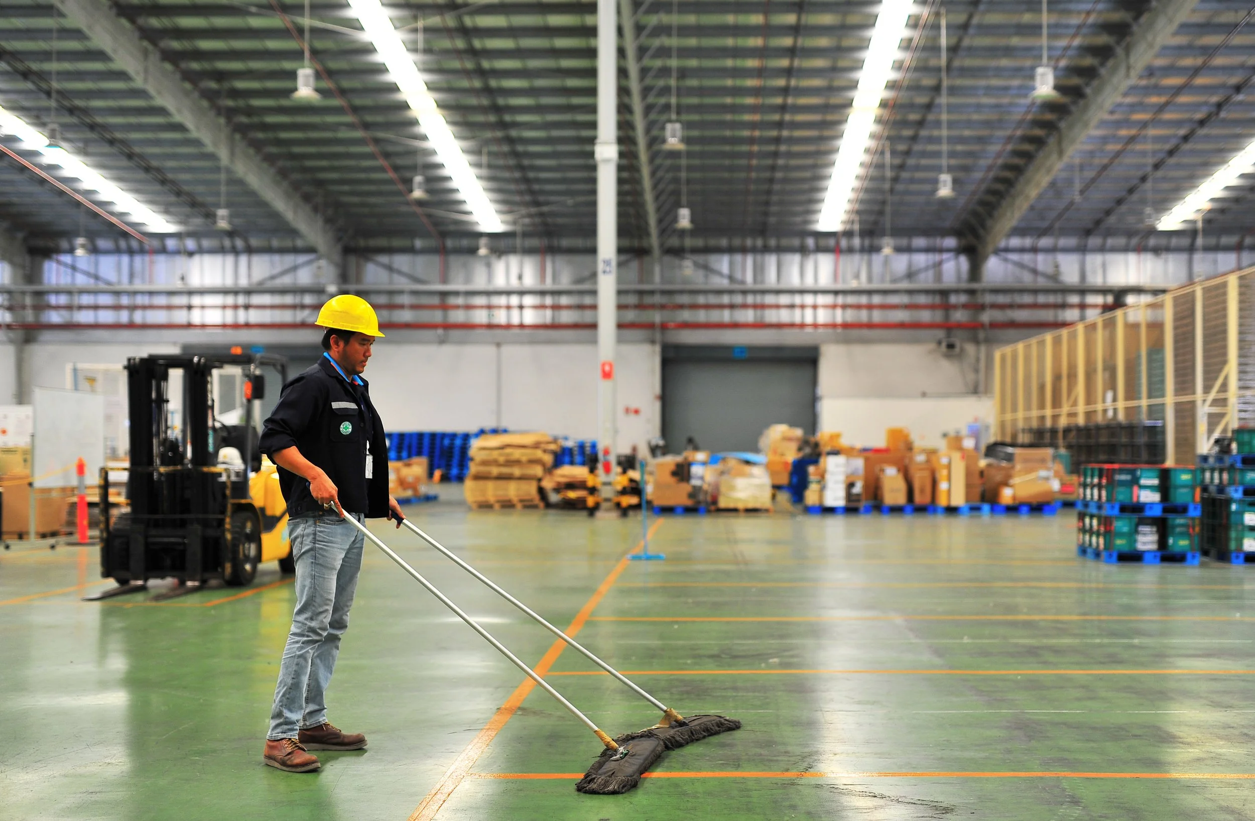 Man in a yellow safety helmet and work clothes cleaning the floor of a warehouse with a mop.