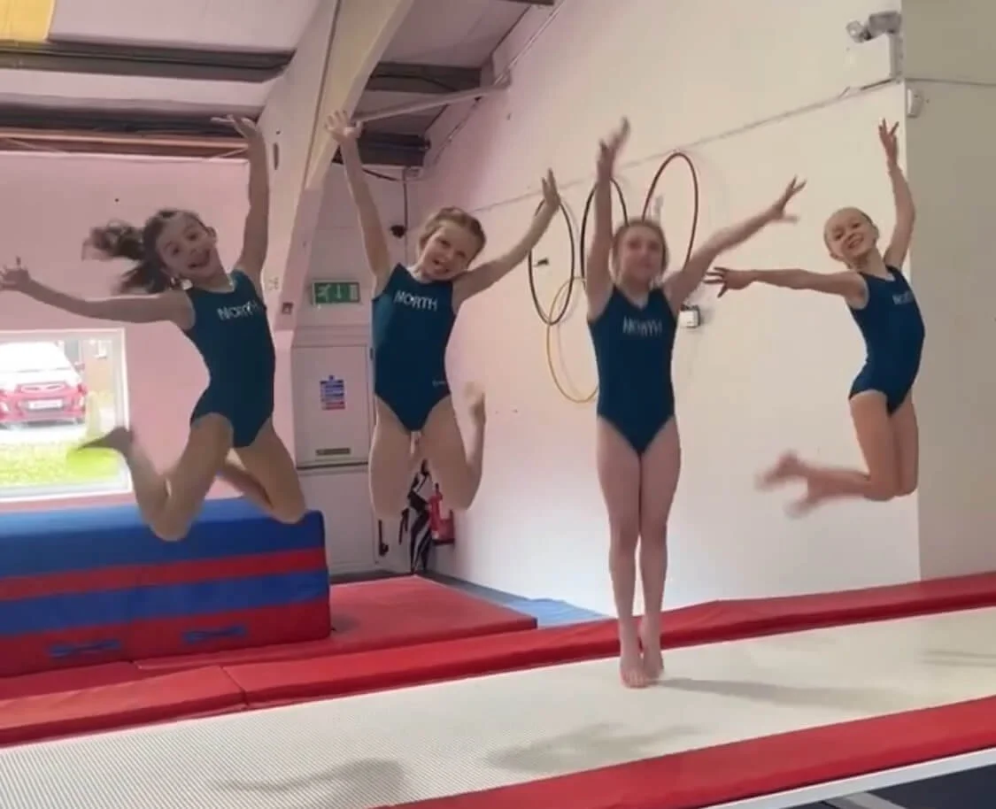 Young girl practicing gymnastics on a balance beam in a gymnasium with her arms raised.