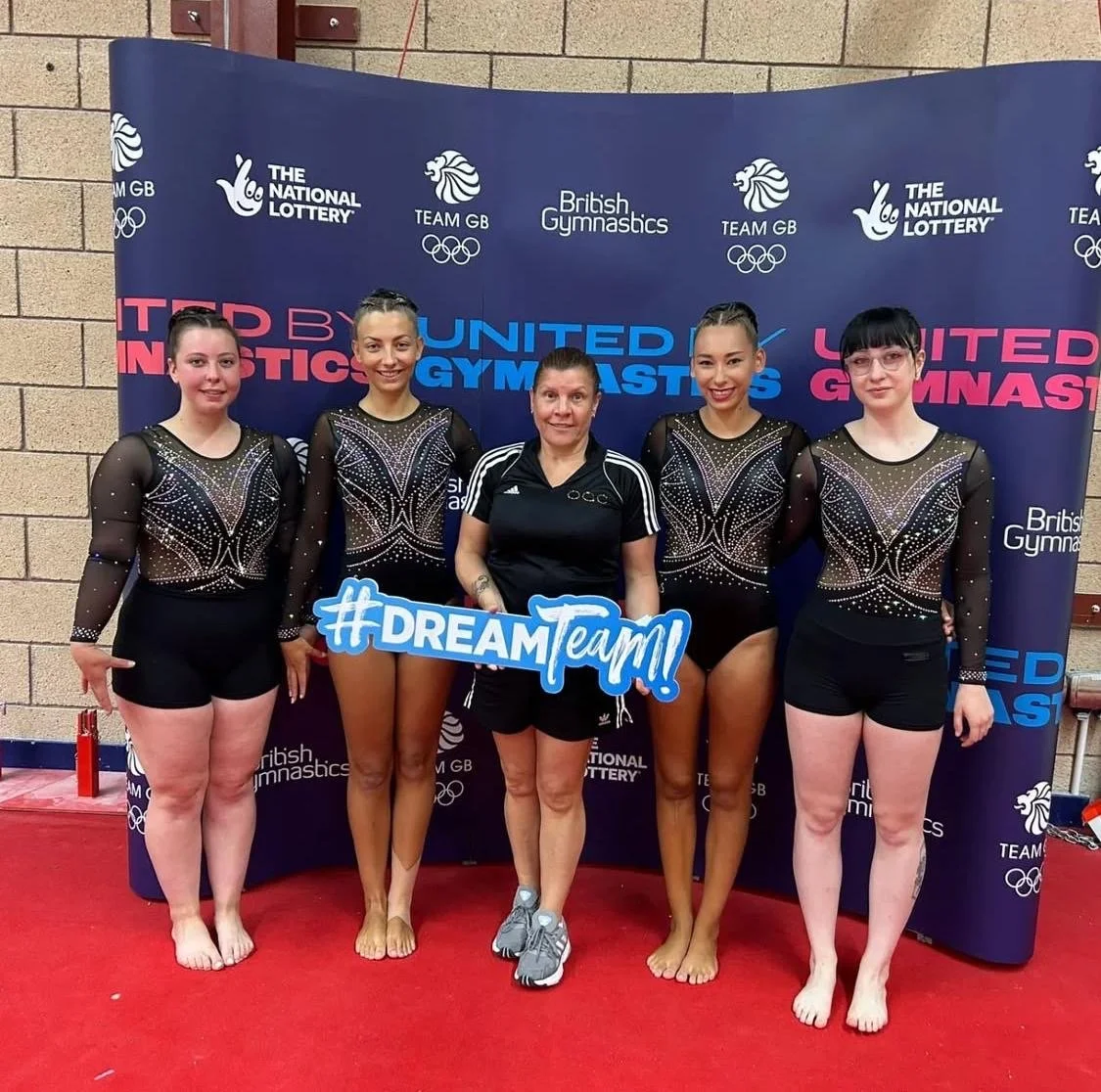 Five young girls sitting and playing on a gymnastics mat in an indoor gym, smiling and laughing.
