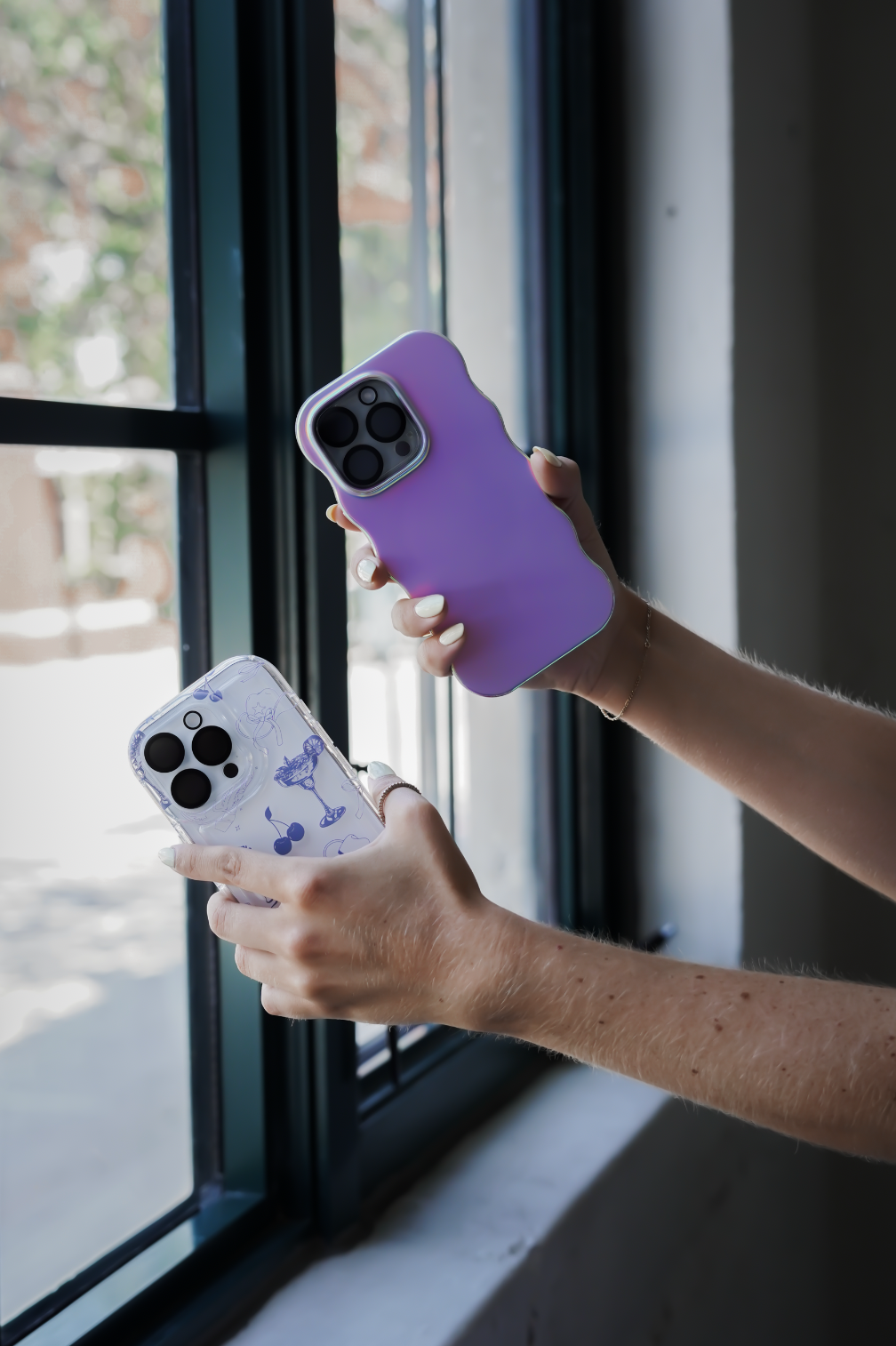 Two hands holding smartphones with colorful cases near a window, with a blurred outdoor scene visible outside.