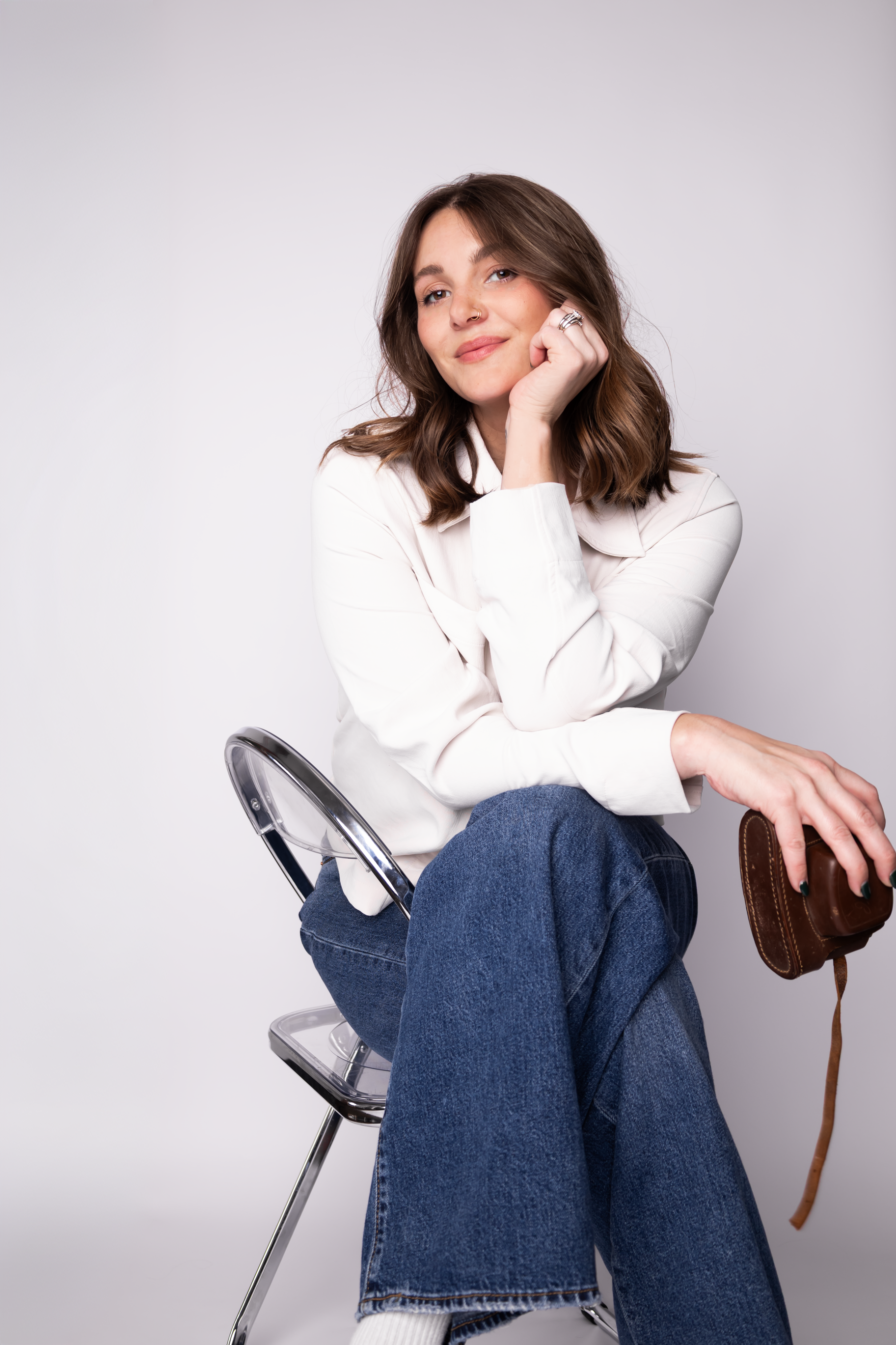 A young woman with brown hair, wearing a white shirt and blue jeans, sitting on a clear chair with her chin resting on her hand, smiling softly, against a plain white background, holding a brown camera in her lap.