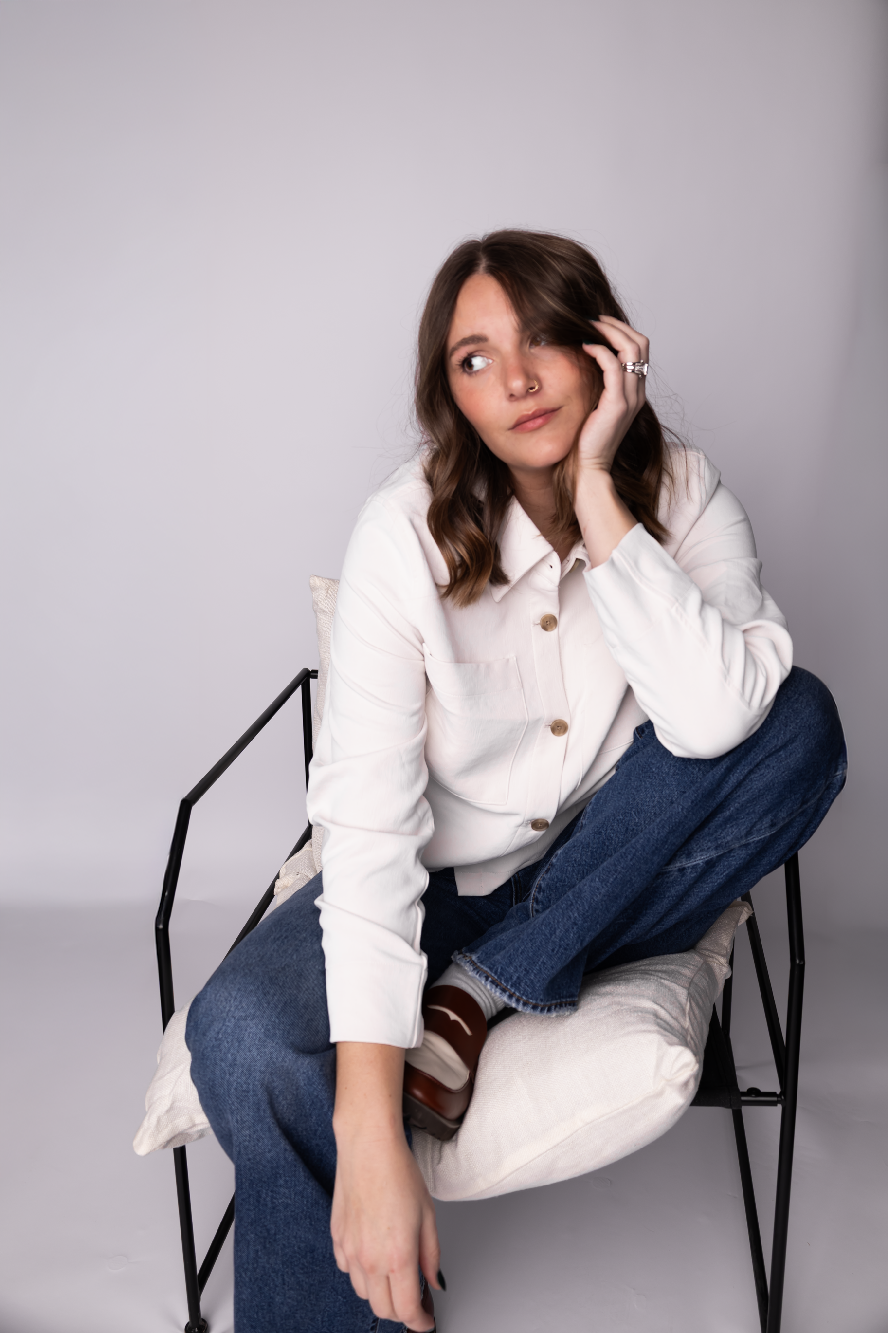 A young woman with brown hair, wearing a white button-up shirt, blue jeans, and brown shoes, sitting on a black chair with a pillow, against a plain gray background.