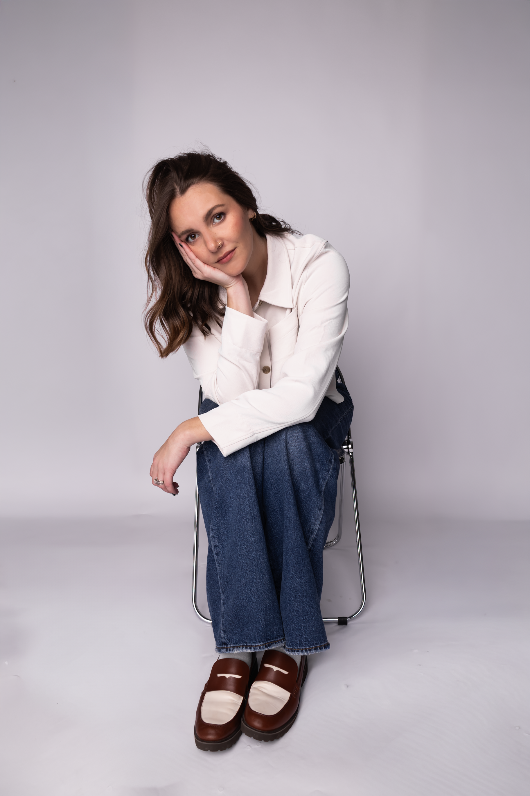 A young woman with shoulder-length brown hair and a nose piercing, sitting on a chair with her head resting on her hand, wearing a white jacket, blue jeans, and brown-and-tan loafers, posing against a plain light gray background.
