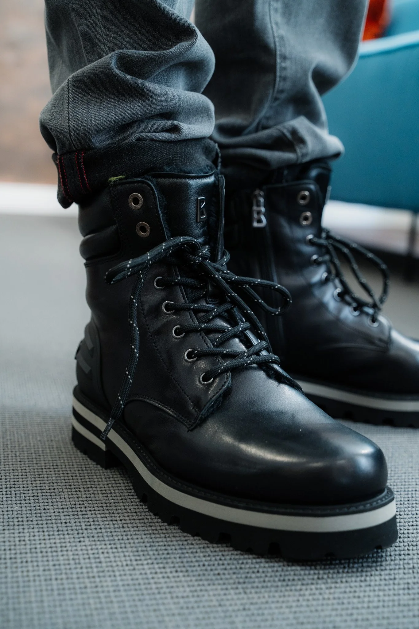 Schwarz Lederstiefel mit dicken Sohlen, getragen von einer Person in grauen Jeans, auf einem grauen Boden.