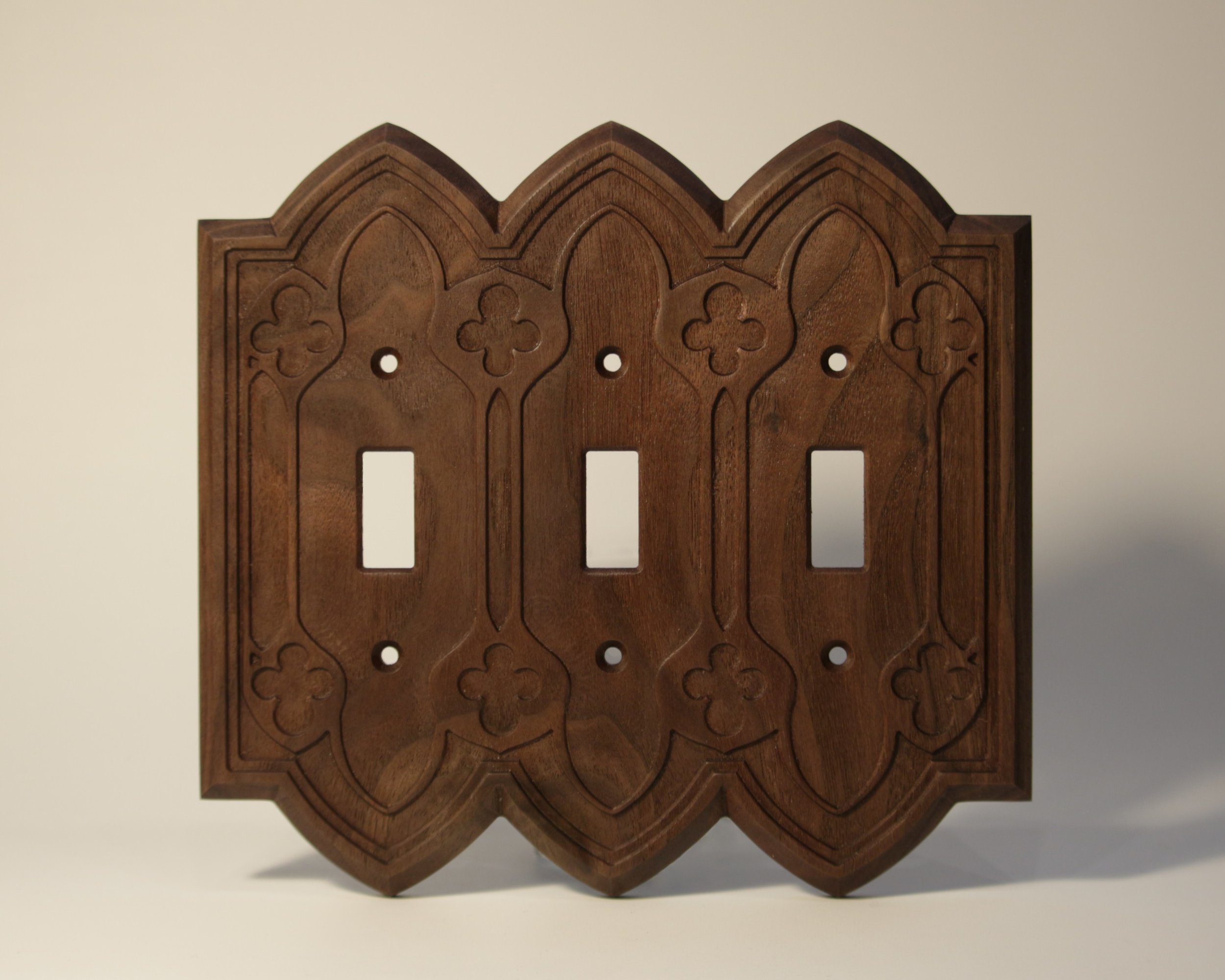 A brown wooden decorative piece with arch-shaped top, intricate carvings, plant motifs, grid cutouts, and small circular holes in the center, set against a plain, light-colored background.