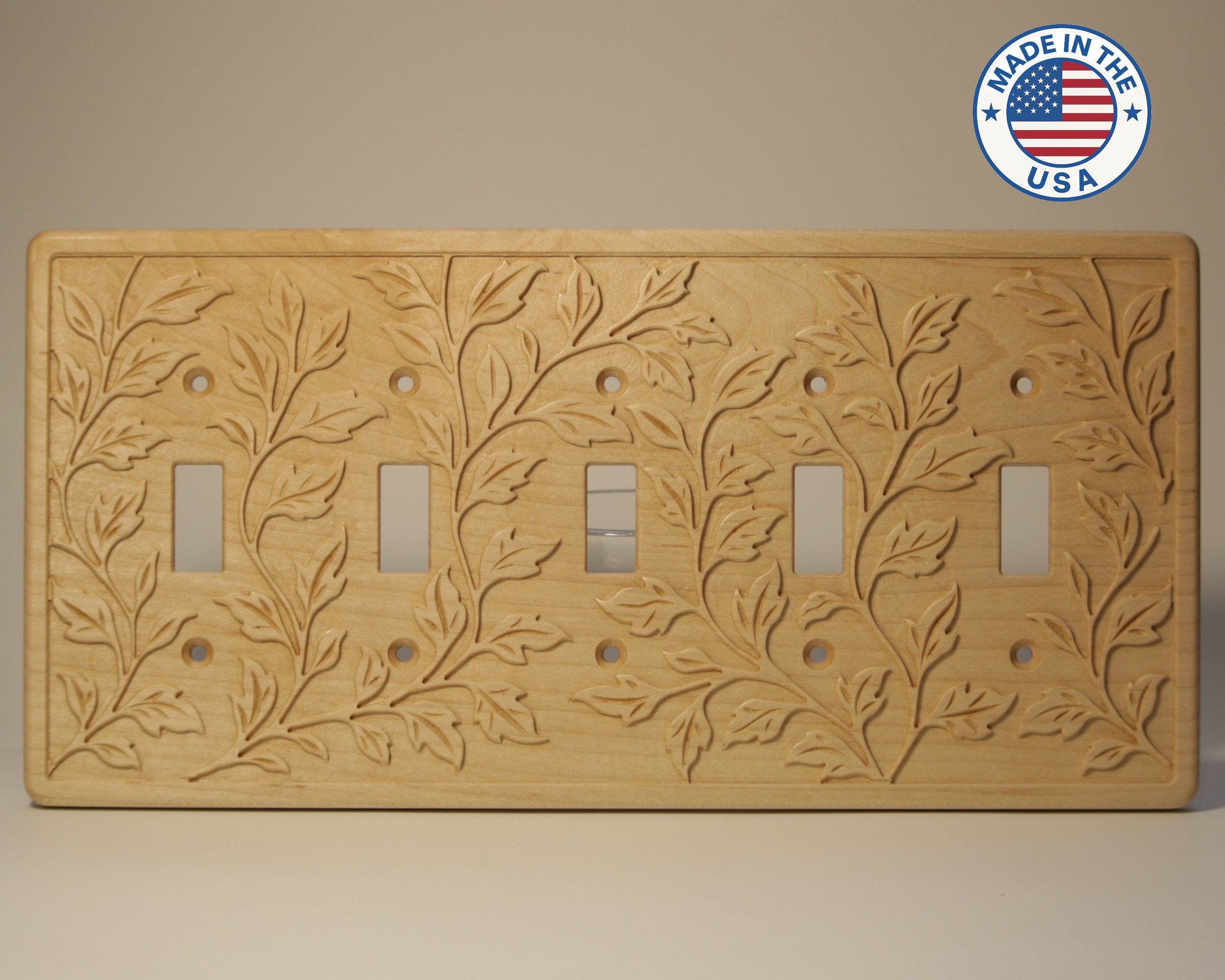 A wooden plaque with intricate leaf and vine carvings and rectangular cutouts, mounted on a gray wall. A round sticker in the upper right corner reads 'Made in the USA' with an American flag inside.