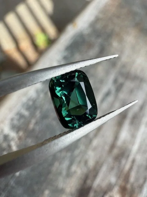A deep green, rectangular gemstone held by tweezers over a rustic wooden surface.