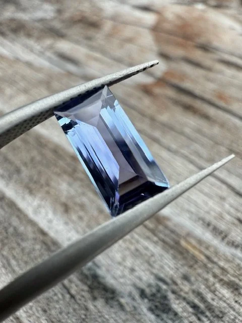 A clear, rectangular, faceted gemstone held by tweezers over a wooden surface.