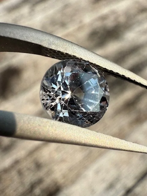 Close-up of a clear, round gemstone, possibly a diamond, held by tweezers against a blurred wooden background.