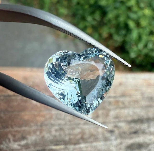A heart-shaped diamond being held with tweezers, outdoors with greenery in the background.