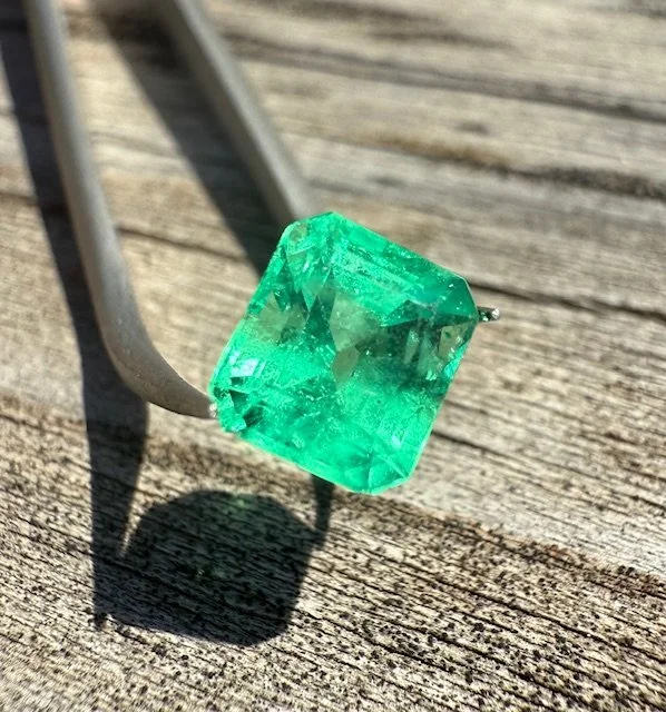 Close-up of a large, rectangular emerald-cut green gemstone resting on a wooden surface with a metal comb partially visible nearby.