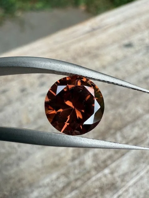A round, reddish-brown gemstone held by silver tweezers outdoors.