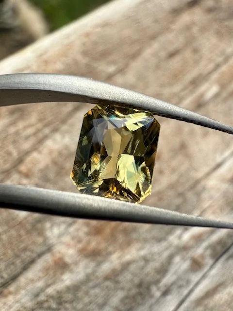 A yellowish-green gemstone held by medical tweezers against a wooden background.