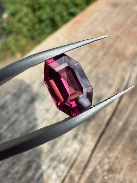A pinkish-purple gemstone held with metal tweezers outdoors on a wooden surface with green foliage in the background.