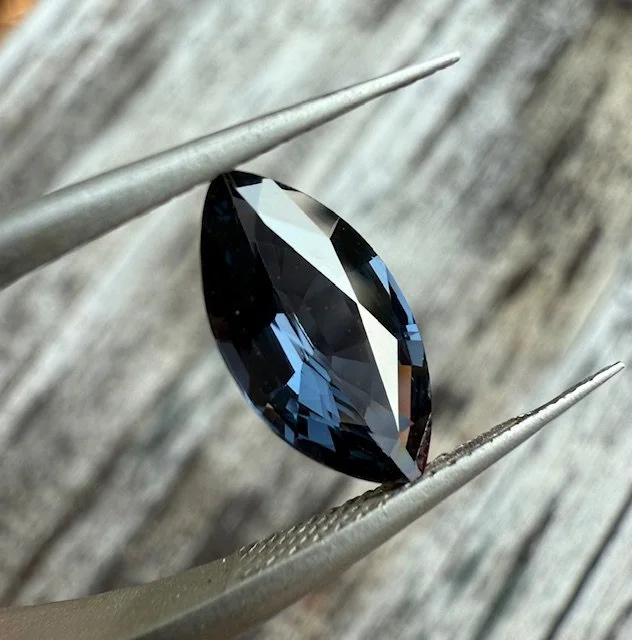 A marquise-cut dark blue gemstone held by metal tweezers against a wooden background.