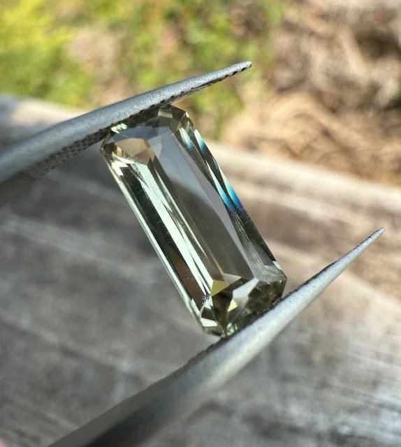 A rectangular cut gemstone being held with tweezers against a blurred outdoor background.