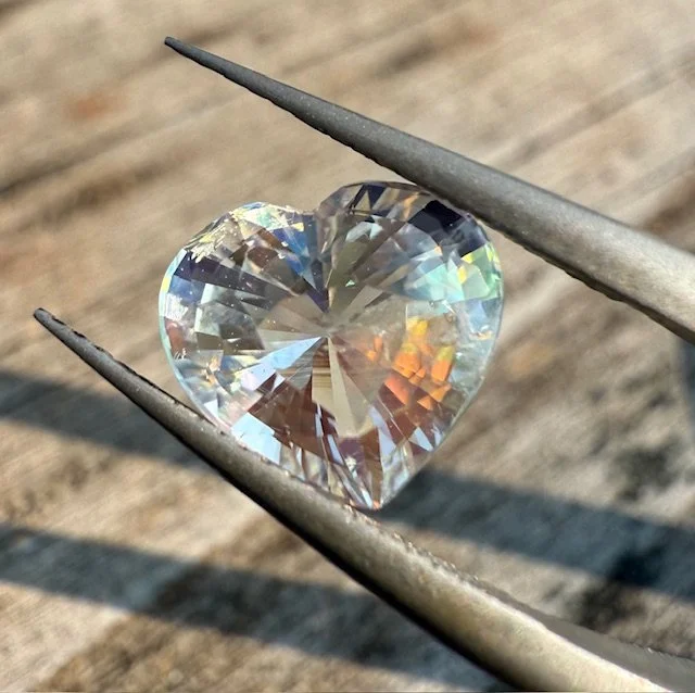 A heart-shaped diamond held by metal tweezers on a wooden surface with sunlight reflection.