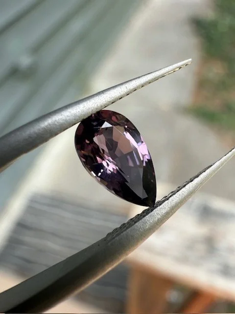 A teardrop-shaped purple gemstone held with jewelry tweezers.
