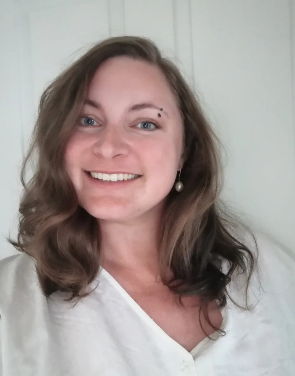 Brunette på hvid baggrund smiler til kameraet, hun bærer hvid skjorte, en synlig perleørering og en sort øjenbrynspiercing