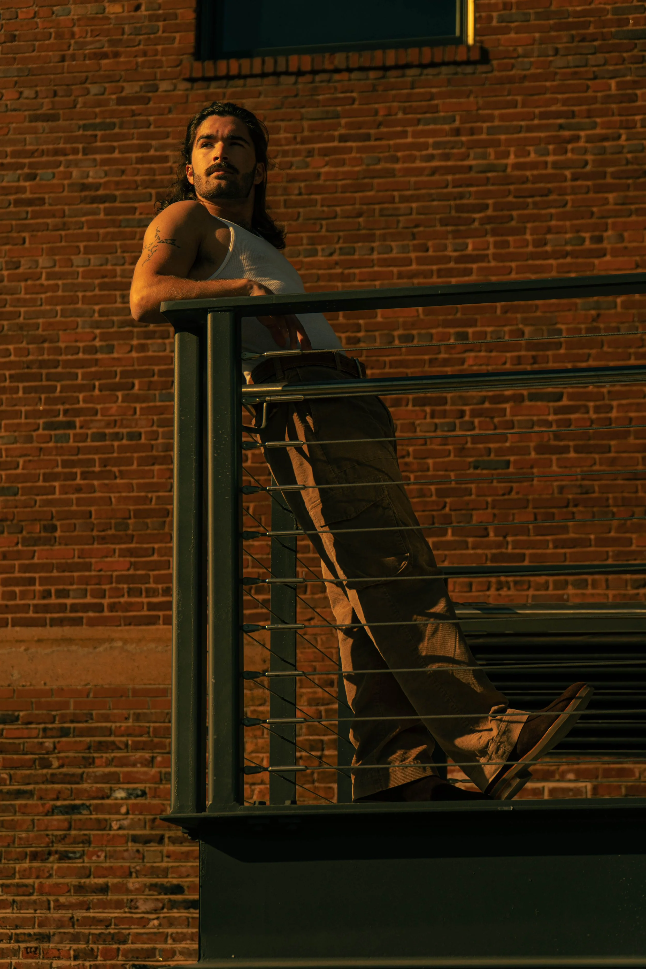 A man with long dark hair, a beard, and tattoos on his arm stands on a balcony with a metal railing, leaning against it while looking off into the distance. He wears a white tank top and dark pants, with a brick building in the background during suns