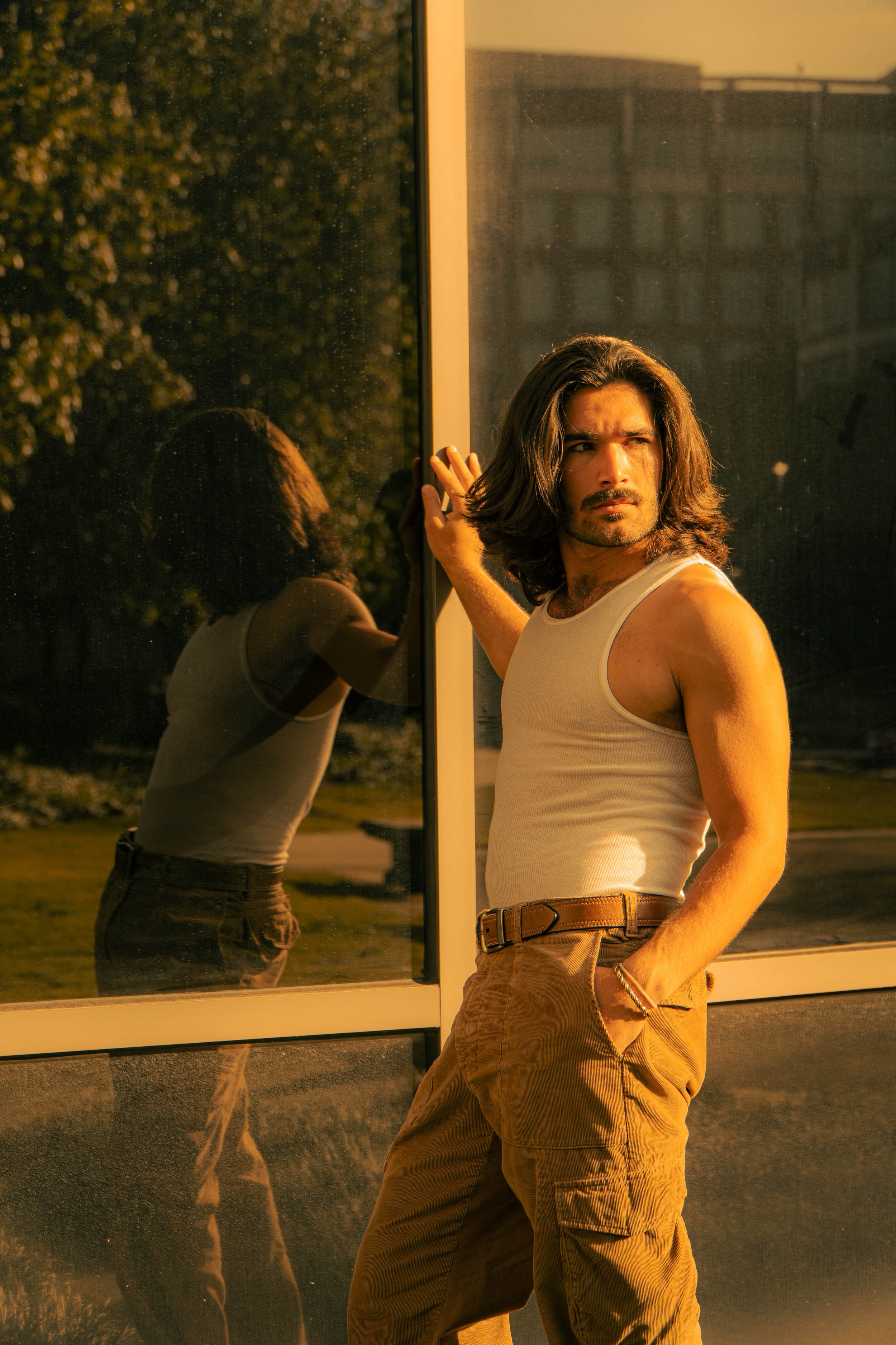 A man with long hair and a mustache, wearing a white tank top and khaki cargo pants, standing outside at sunset, leaning against a glass window which reflects his image.
