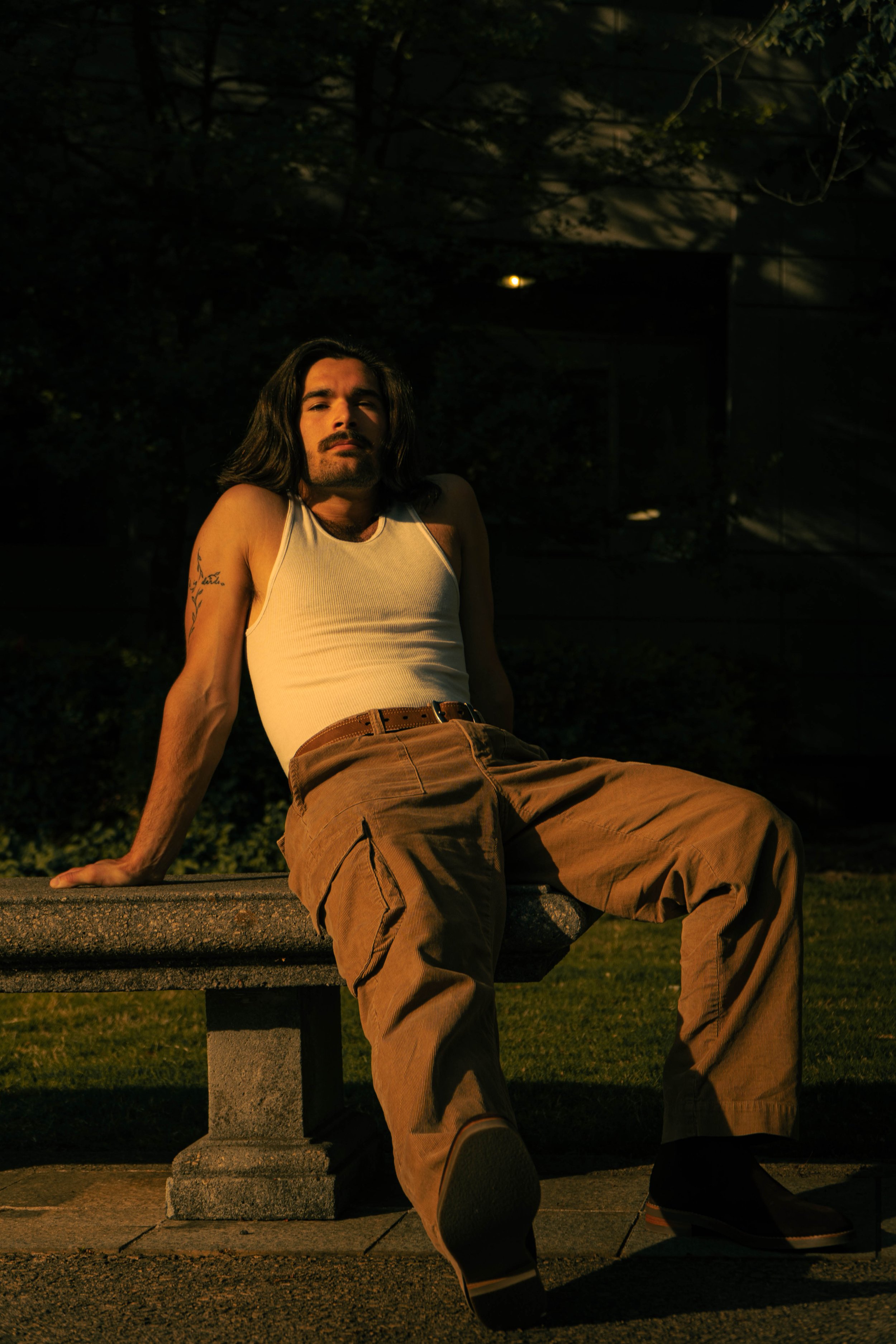 A man with long dark hair and a mustache sitting on a park bench at night. He is wearing a white tank top and brown pants, with one leg extended forward and his hand resting on the bench. The background features dark trees and dim lighting.
