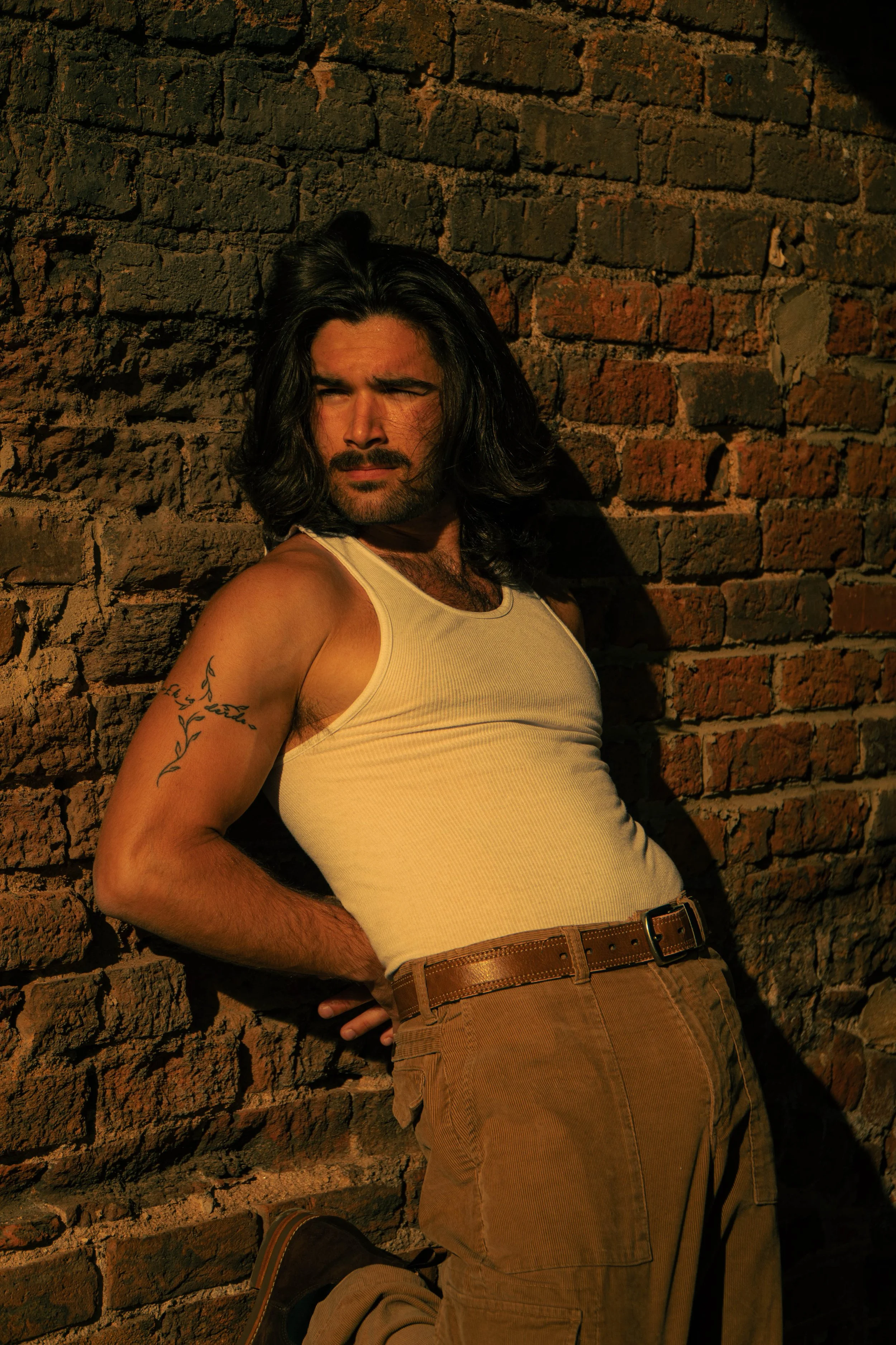 A man with long dark hair and a beard leaning against a brick wall, wearing a white tank top and brown pants, with the sun illuminating his face.
