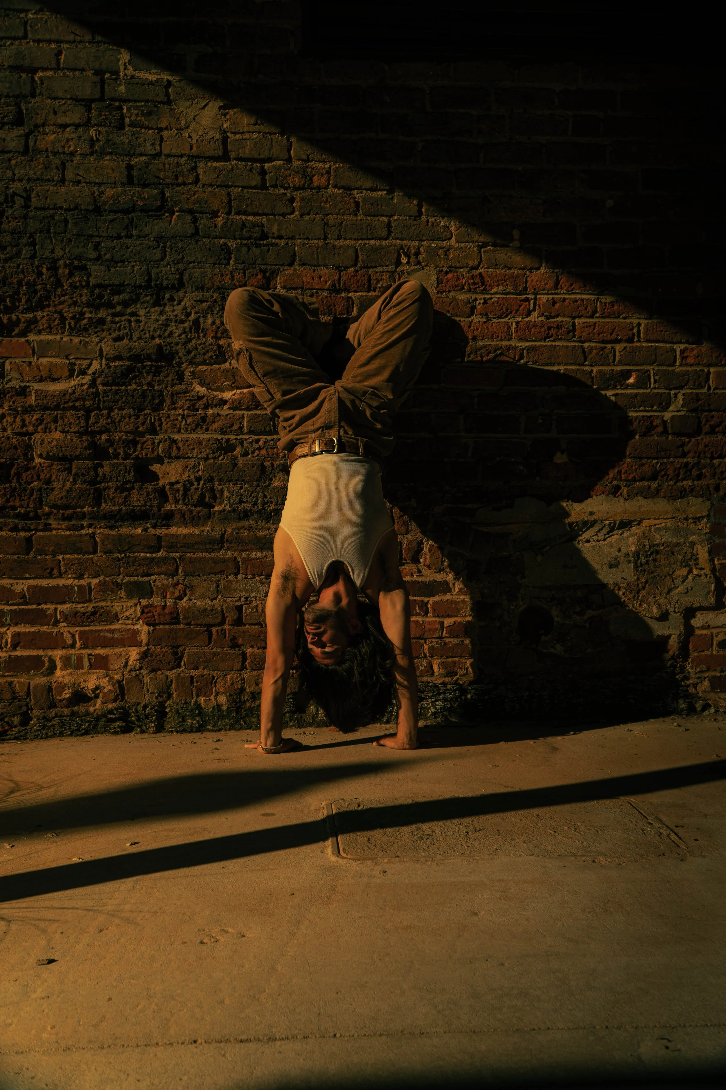 Person performing a handstand against a brick wall during the evening, casting a shadow on the ground.