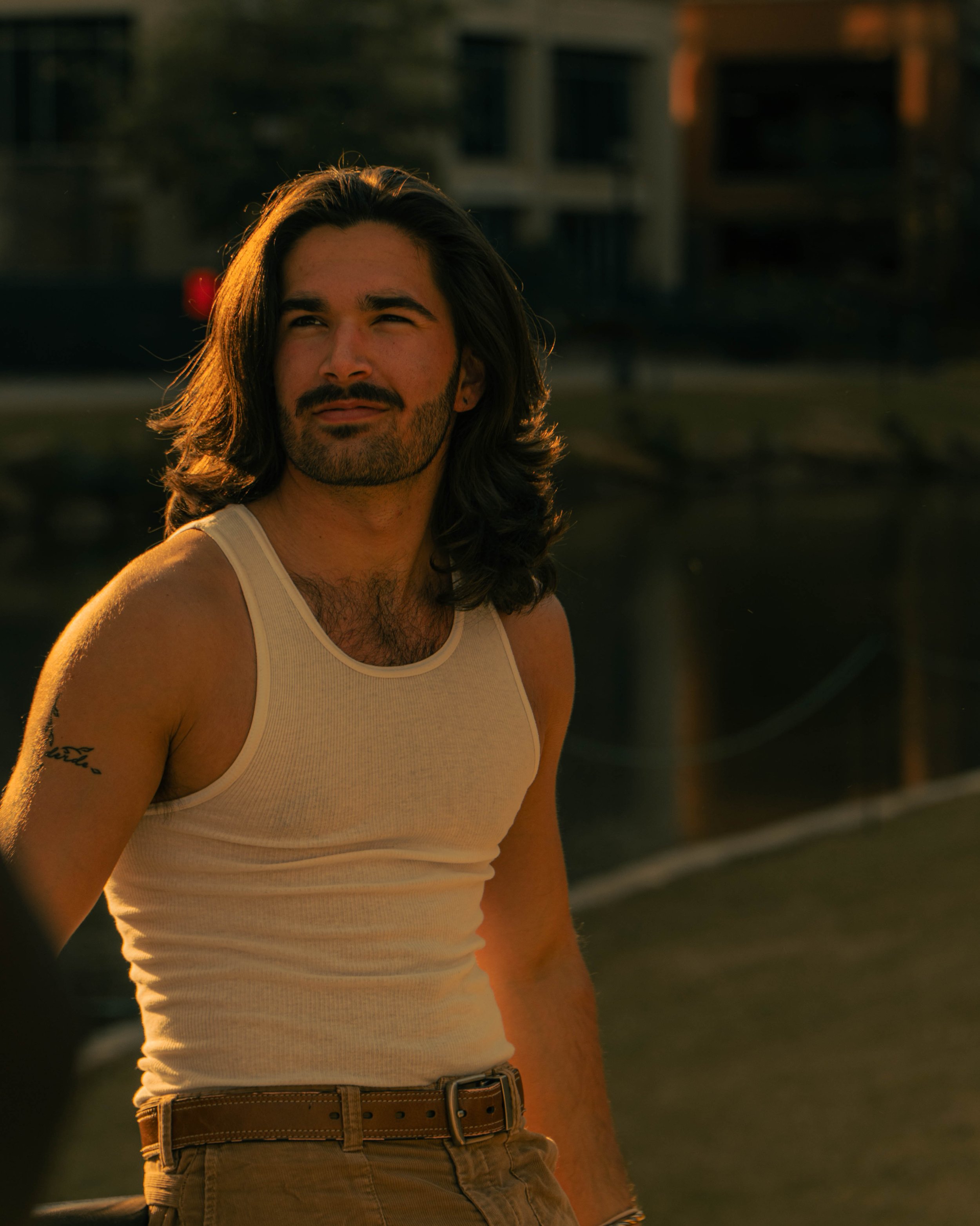 Close-up of a man with long dark hair, a beard, and mustache, wearing a white tank top and khaki pants, outdoors during sunset.