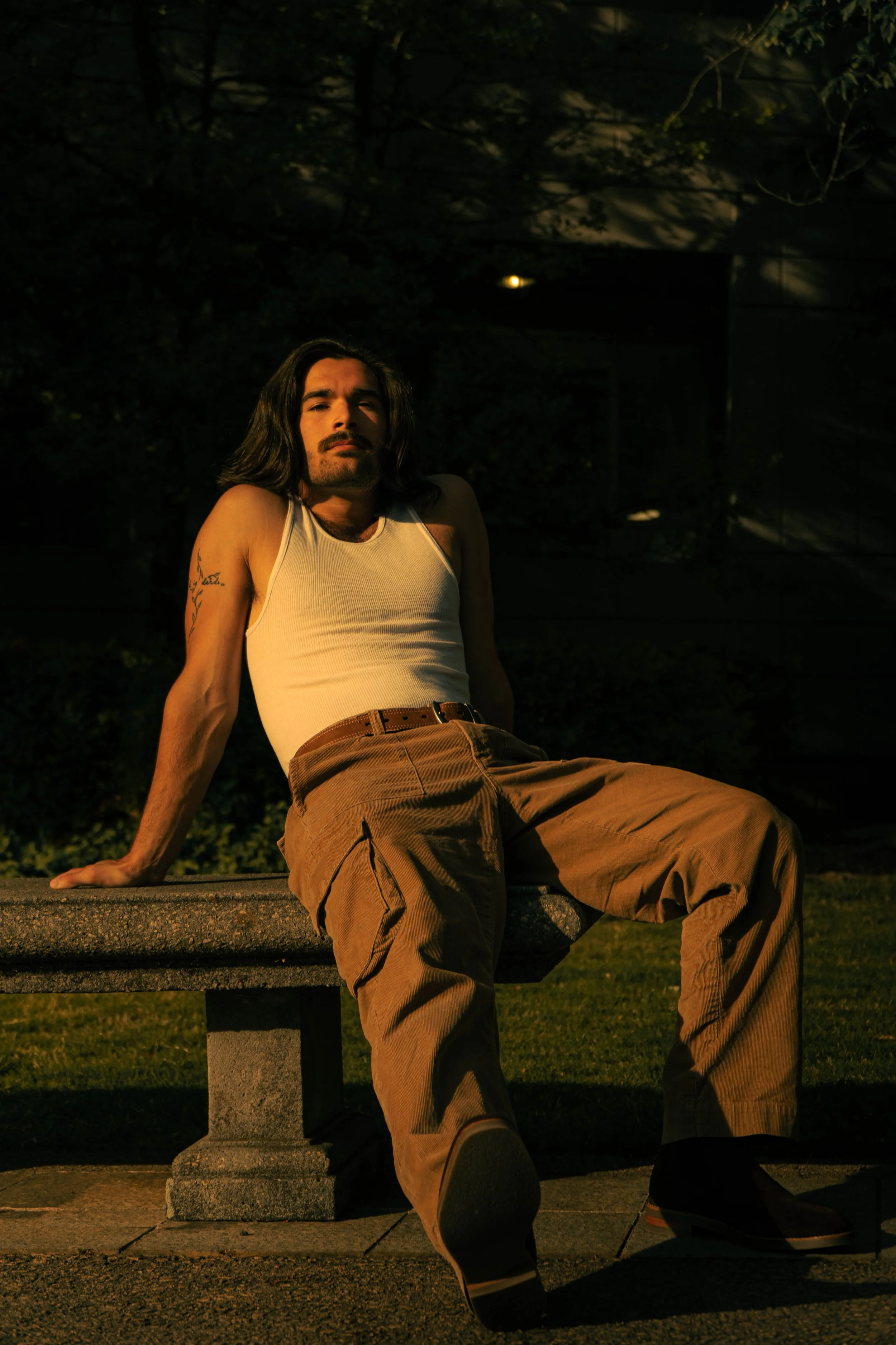 A man with long dark hair, a mustache, and beard, wearing a white tank top and brown cargo pants, sitting on a concrete bench at night with trees and a dark building in the background.