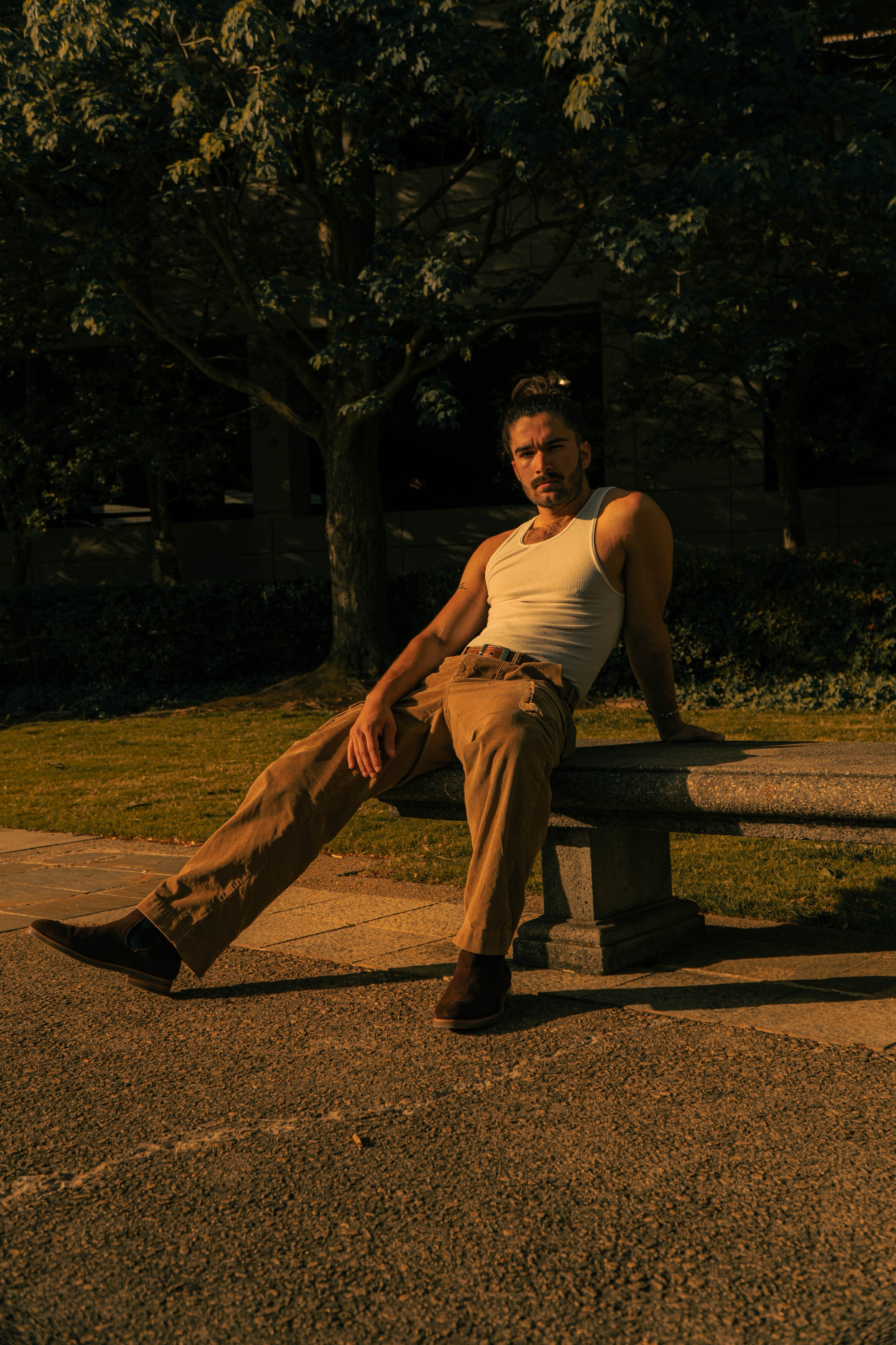A young man with dark hair and a beard, wearing a tan tank top and tan pants, sitting on a park bench at sunset with trees in the background.