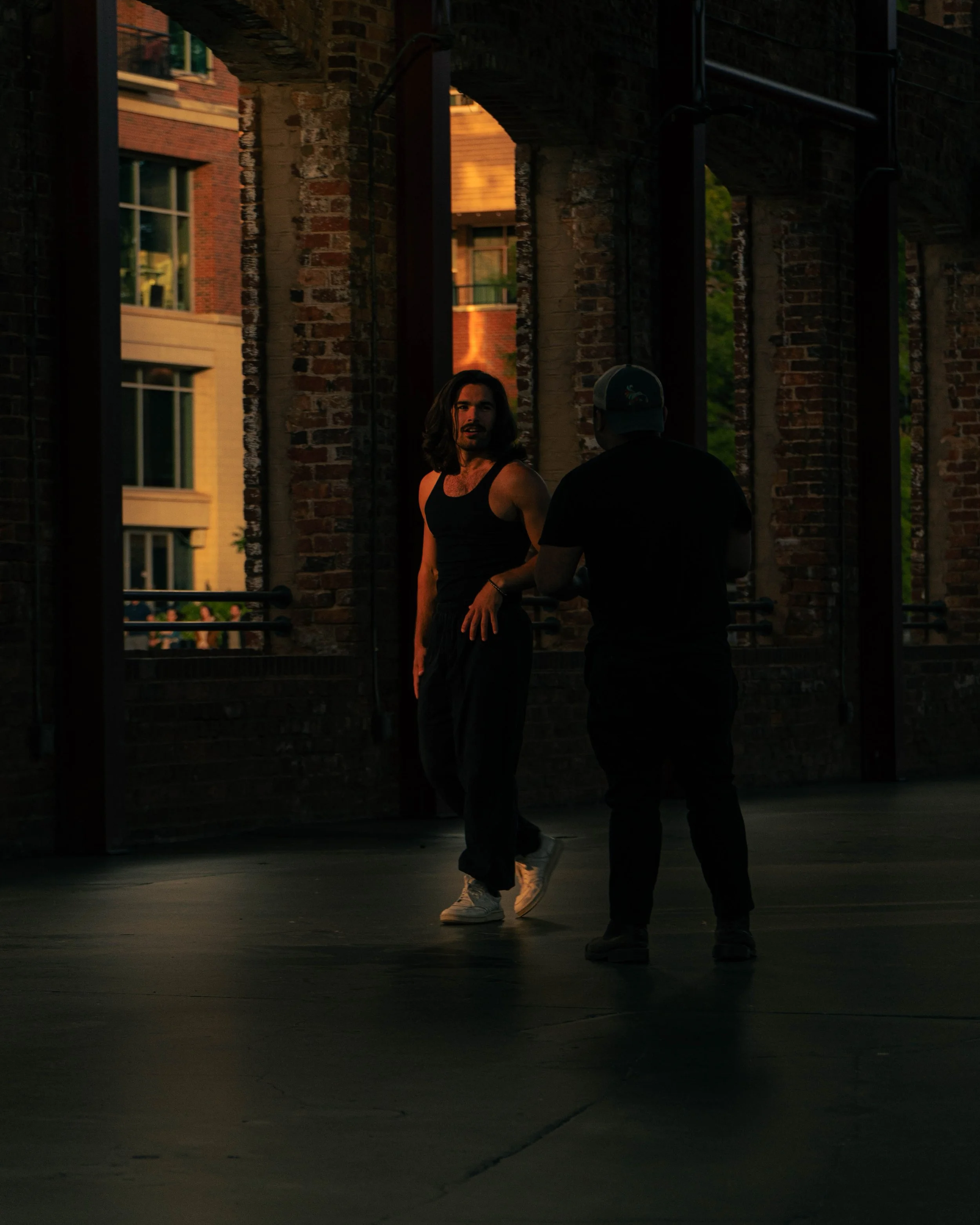 A man with long hair and a beard wearing a black tank top and black pants standing on the street at dusk, talking to a person wearing a baseball cap and black clothing. The background features brick buildings and windows, with some reflections and gr